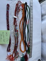 Nine strands of new glass beads with tags, arranged next to a ruler for scale. Colors include clear, orange, green, red, black, and multicolored. Various bead shapes and sizes visible.