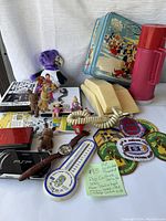 Lot overview showing handheld consoles, toys, VHS, thermos, phone, patches, and photo cards.