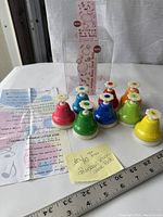 Set of multi-colored vintage mini desk bells with floral key labels arranged on a white surface alongside the instructional paper sheet and original packaging.