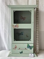 Front view of the light green painted wooden cabinet with wire-backed door featuring butterfly decals and drawer at the bottom.