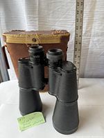 Vintage Carl Wetzlar Falcon heavy binoculars standing in front of a very worn, torn brown carrying case with fabric lining and snap button flap.