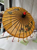 Top view of a vintage parasol showing yellow fabric canopy with hand-painted flowers and birds, black wooden ribs, and decorative tassels at each rib tip, with red pom-pom at the finial.