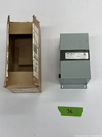 Photo showing the transformer with its original cardboard box packaging, the transformer is gray metal with mounting flanges and a label indicating specifications.