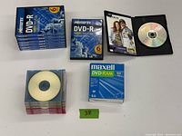 Photo showing stacked blue-packaged Memorex 6-pack DVD-R and individual DVD-R CD cases, Maxell DVD-RAM 5-pack spindle, and loose individual disks.