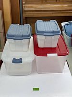 Four clear plastic storage bins stacked on table. Two Rubbermaid bins with gray lids on top of two larger bins, one with a red lid and one with a white lid and gray latch.