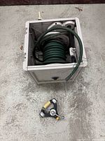 Top view of square plastic hose reel unit containing coiled green garden hose, with metal tripod sprinkler placed beside it on floor.