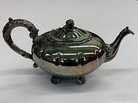Side-angle view of silver plated tea pot showing ornate handle, lid with knob, and curved spout, reflective surface with some tarnish.