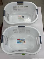 Top view photo showing two white Rubbermaid laundry baskets with gray handles placed on a table. Each basket has ventilation holes on the sidewalls and a product label on the inside bottom.