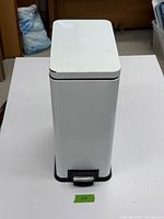 Front view of white step-on waste can showing pedal and rectangular shape with some surface marks.