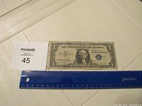 1957B Blue Seal $2 dollar bill with a plastic ruler for scale and a lot number card beside it on a white surface.