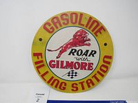 Front view of the round metal Gilmore Gasoline sign with lion logo and text 'Roar with Gilmore' and 'Gasoline Filling Station' in red lettering on yellow and white background.