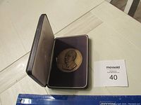 Closed display case with visible bronze medal inside, showing the relief portrait of Lyndon B Johnson and a star border, on a wooden table alongside a ruler for scale.