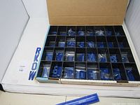 Image of open box showing the blue plastic letter and number stencils organized in compartmentalized plastic tray inside a cardboard box, along with ruler for scale.