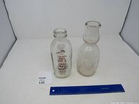 Two vintage glass milk bottles, one with printed Farm Fresh Maine Milk branding and one plain clear glass bottle, placed next to a blue ruler for scale.