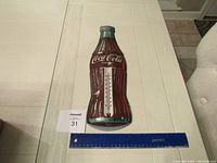 Full view of the Coca-Cola bottle shaped metal thermometer lying flat next to a measuring ruler showing scale approximately 24 inches long.