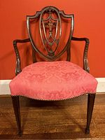 Front view of carved mahogany chair against red wall showing shield-shaped back, scrolled arms, and upholstered seat.