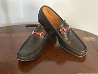 Pair of black leather Gucci loafers angled on wooden surface, showing red-green webbing and horsebit detail on vamp