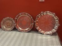 Three vintage silver plated serving trays displayed side by side showing ornate edges and surface wear