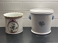 Two ceramic decorative pots side by side; left pot white with scalloped gold rim and painted bird, right pot plain white with small floral design and lion head handles