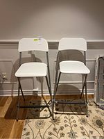 Front view of two white folding bar stools with metal frames and composite seats and backs, placed side by side on a patterned rug.