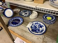 Wide view of multiple blue ware plates on wooden shelf showing various blue patterned plates in different shapes and sizes including some with floral designs and a set of small white plates with blue rims.