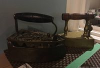 Photo showing two brass sad irons with wooden handles on a table, dark and light iron visible