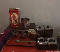 Photo showing the vintage canisters, bread box, and two collector tins on a red surface against a gray wall.