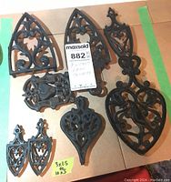 Full set of eight black cast iron trivets arranged on cardboard, showing different shapes and sizes, with MaxSold tag for identification.