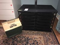 Black metal organizer with 12 mesh pull out trays placed on a carpet with a vintage Little Playmate cooler beside it.