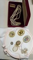 Photo showing the triple string of pearls in a dark red velvet-lined box alongside assorted vintage costume jewelry, including brooches, a locket, and ornamental pins, with a mini guarantee booklet placed above.