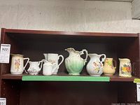 Eight porcelain creamers and pitchers arranged on a shelf, showing a variety of styles, colors, and floral decorations.