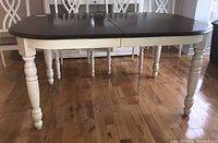 Full view of wood dining table with dark brown top, white turned legs, and leaf inserted in center.