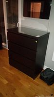 Front view of black manufactured wood 5 drawer dresser with smooth top and closed drawers placed on wooden floor.
