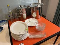 Wide shot of stainless steel pots with lids, glass bottles, white Lubina dinner plates, clam shell shaped glass dishes, glass bowl and rectangular glass dish on table.