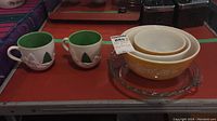 Overall lot showing the two Starbucks Christmas mugs, three Pyrex nesting bowls, and clear pie plate on a shelf