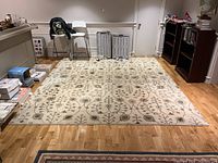Photo showing the full rug spread on a hardwood floor in a room with light walls, bordered with shelves, chairs and boxes around it.
