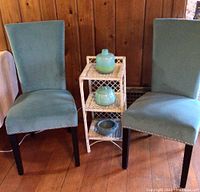Two light blue velvet high back chairs flanking a white wicker 3-tier shelf with three blue pottery pieces displayed.