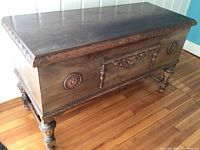 Full view of antique cedar trunk showing carved front panel with floral and medallion details and turned legs.