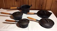 All five cast iron pots and pans arranged on a white cloth background with wooden walls in background. Features wooden handles and four lids, one lid designed to fit two pieces.