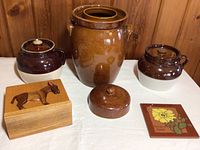 Overview photo of all items including large fermentation crock, two smaller crocks, ceramic pot, wooden donkey box, and ceramic tile on a white cloth background against wooden panel wall.