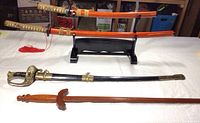 Two orange-lacquered Japanese Tachi blades on black wooden stand, beside wooden sword and US saber