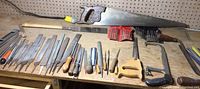 Wide view of multiple metal files, hand saw with wooden handle, chisels, and coping saw on wood shelf