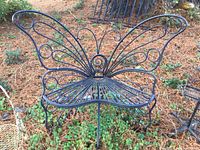 Iron butterfly-shaped metal bench with curved wire design shown outdoors on grass and pine needles.