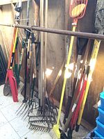 Wide view showing multiple long handled garden tools including rakes, shovels, and a broom in a shed against a wooden wall.