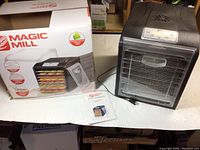 Photo of Magic Mill food dehydrator with power cord, clear front door, and stainless steel interior shelves visible. User manual placed next to the dehydrator. The dehydrator appears clean and lightly used.