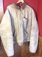 Beige Carhartt jacket hanging on wall, showing front with zipper, pockets, and corduroy collar.