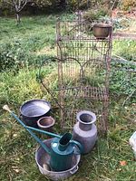 Overall lot showing weathered metal foldable garden shelf with heart-shaped hanging plant holders, green watering can, metal milk can, and various pots on grass.