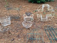 Several metal planting baskets, plant stands, and trellises arranged outdoors on pine straw-covered ground showing various shapes and colors including white, green, and rust.