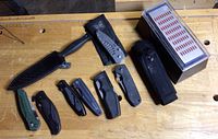 All items laid out on wooden workbench: folding knives, dagger with sheath, nylon sheath and sharpening stone