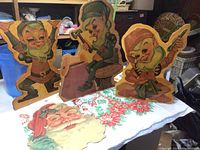 Three painted wood elf figures and a Christmas-themed vinyl tablecloth with Santa face and poinsettia patterns.